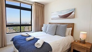 Ein Hotelzimmer mit einem großen Bett, einem Nachttisch mit Lampe, einem Bild von einem Boot an der Wand und Blick auf die Stadt durch das Fenster.