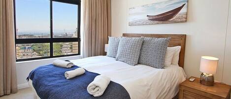 A hotel room with a large bed, a nightstand with a lamp, a painting of a boat on the wall, and a view of the city through the window.