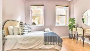 A bedroom with a bed, two windows, a desk, a chair, and a potted plant. - The Piazza (Cape Town)