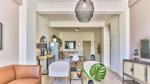 A spacious living room with a sofa, chairs, a coffee table, and a large plant. A kitchen is visible in the background with a dining table and chairs. - The Piazza (Cape Town)