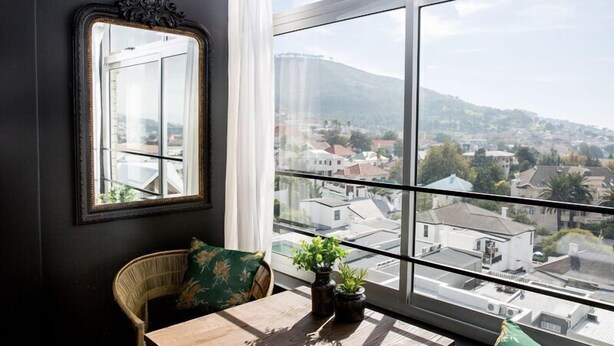 Ein Zimmer mit großem Fenster und Blick auf eine Stadt und Hügel. An der Wand hängt ein Spiegel und ein Tisch mit zwei Topfpflanzen.