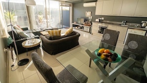 A modern living room with a sofa, a glass coffee table, and a dining area with chairs and a fruit bowl. - The Legacy II (Cape Town)