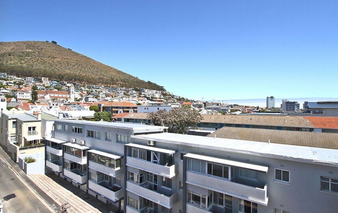 A coastal town with modern apartment buildings, a prominent hill in the background, and a clear blue sky.