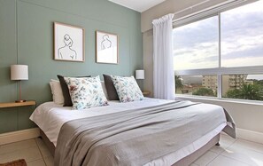 Een slaapkamer met een groot bed, twee aan de muur bevestigde fotolijstjes, een raam met gordijnen en uitzicht op gebouwen.