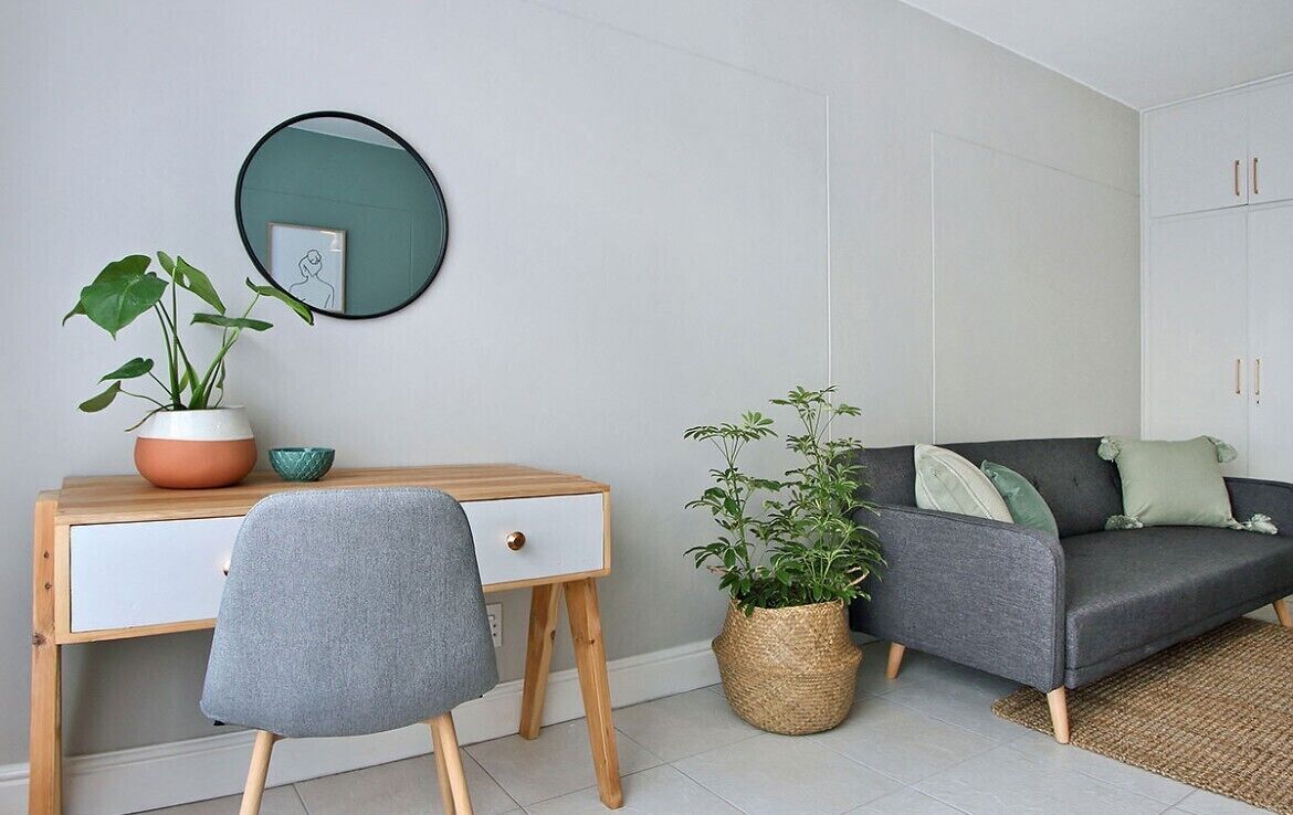 Een moderne zithoek met een grijze bank, een houten bureau met stoel, een ronde spiegel en twee potplanten.