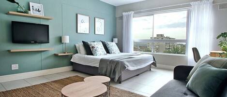 A modern bedroom with a bed, a TV, a sofa, and a window with a city view.