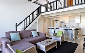 A modern living room with a sofa, coffee table, and wicker chairs. A staircase leads to a kitchen with a bar area.