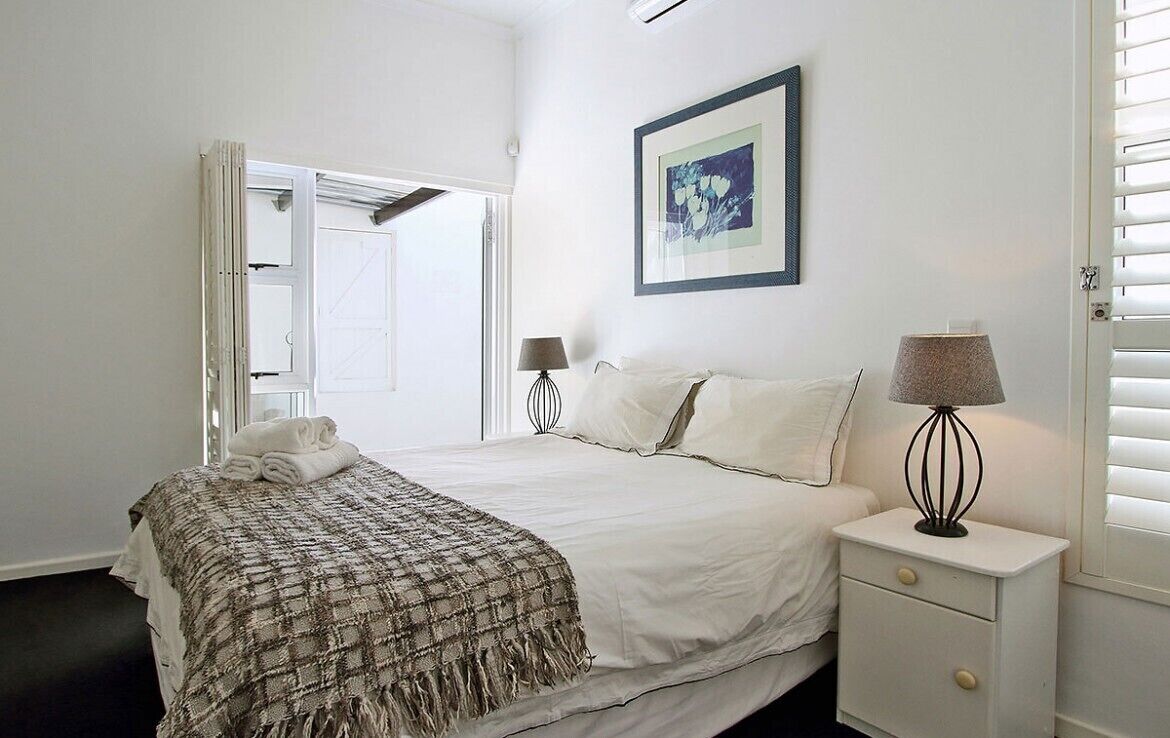 A bedroom with a bed, a bedside table, a lamp, a framed picture on the wall, and a window with blinds.
