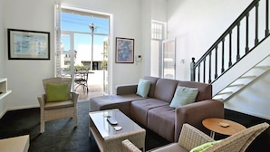 Ein modernes Wohnzimmer mit einem braunen Sofa, Korbstühlen und einem Glastisch. Eine Treppe ist rechts zu sehen.