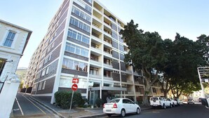A multi-story residential building with a brick facade, multiple balconies, and a parking area with several cars.