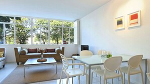 A modern living room with a sofa, coffee table, and dining table with chairs. Large windows provide natural light and a view of trees.