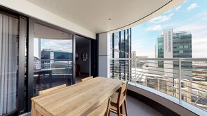 Un balcón moderno con una mesa y sillas de madera, ventanas amplias que ofrecen vistas a la ciudad y una puerta que da a una habitación contigua.