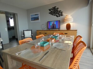 Un comedor con una mesa de madera dispuesta para seis personas, sillas anaranjadas, un televisor en la pared y un adorno decorativo encima del televisor.