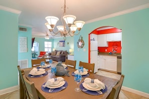 A dining area with a wooden table set for six, a chandelier, and a kitchen in the background.