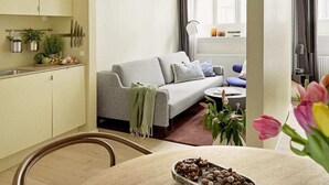 A modern living room with a grey sofa, a small table, and a kitchen area with cabinets and a plant. - One-Bedroom Apartment for 3 (København)
