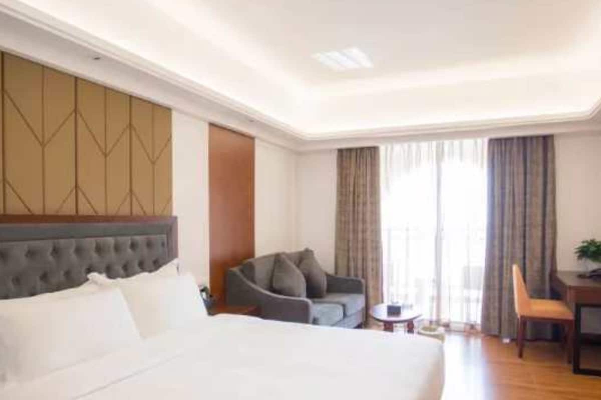 A hotel room with a large bed, a grey sofa, a small table, and a chair.