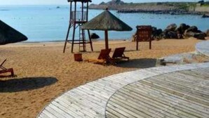 Una playa con muelle de madera, sillas de descanso, una torre de salvavidas y una sombrilla de paja.