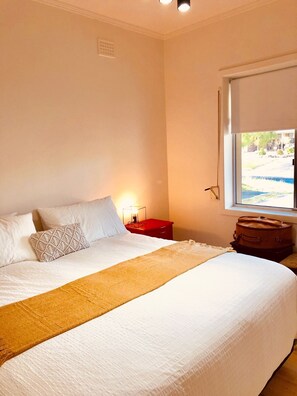 A neatly made bed with a white comforter and a brown throw blanket, a red bedside table with a lamp, and a window with a view of greenery.