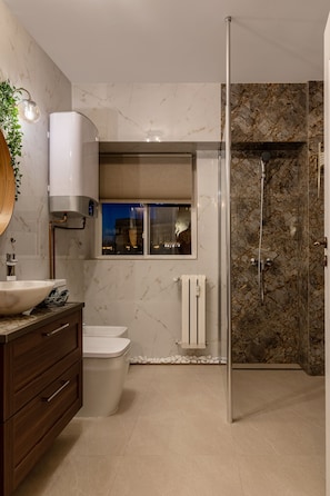 A modern bathroom with a marble wall, a white sink, a toilet, a shower with a glass door, and a window with a view of the city at night.