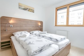 A bedroom with a wooden headboard, two beds with white bedding, a small bedside table, a wall-mounted light, a window with blinds, and a radiator.