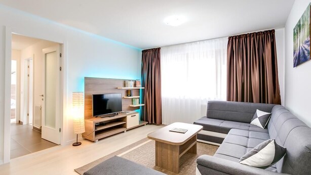 Ein modernes Wohnzimmer mit grauem Sofa, einem Holztisch, einem Flachbildfernseher auf einem Holzständer und einem großen Fenster mit braunen Vorhängen.