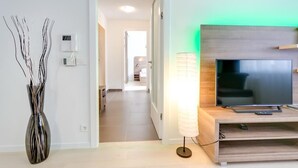 Ein modernes Wohnzimmer mit Flachbildfernseher auf einem Holzständer, einer Stehlampe und einer Wandvase mit Ästen.