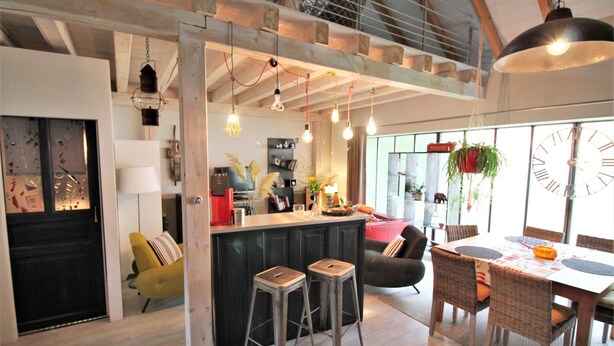 Una cocina moderna con barra, vigas de madera, lámparas colgantes y zona de comedor con un reloj.