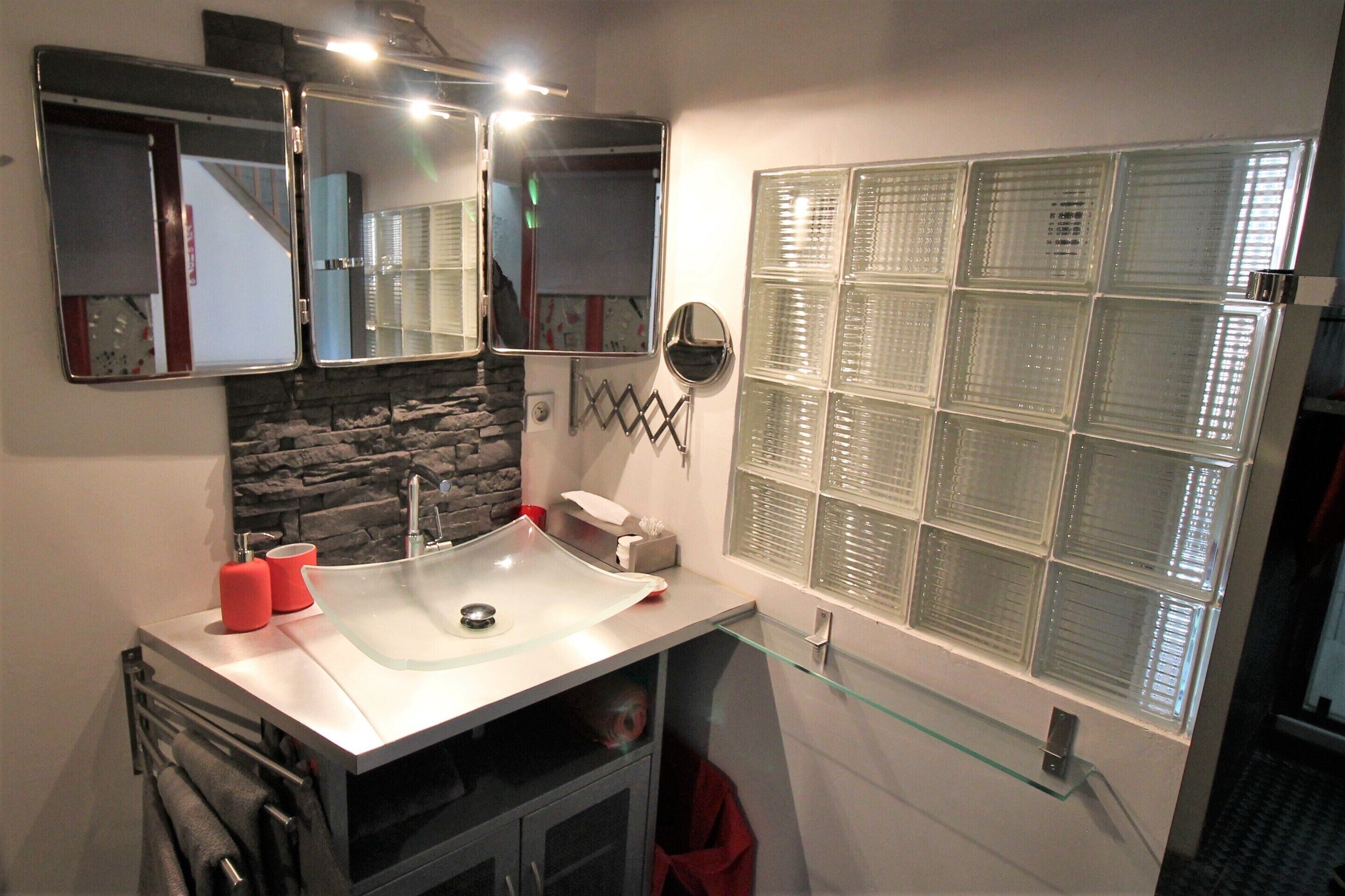 A bathroom with a glass sink, two mirrors, a stone accent wall, and a window with blinds.