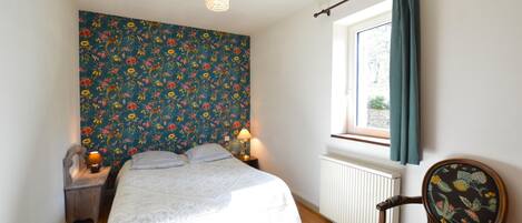 Et soveværelse med blomstret tapet, en seng med hvid dyne, et trænatbord med en lampe, en stol med blomstret betræk og et vindue med gardiner.
