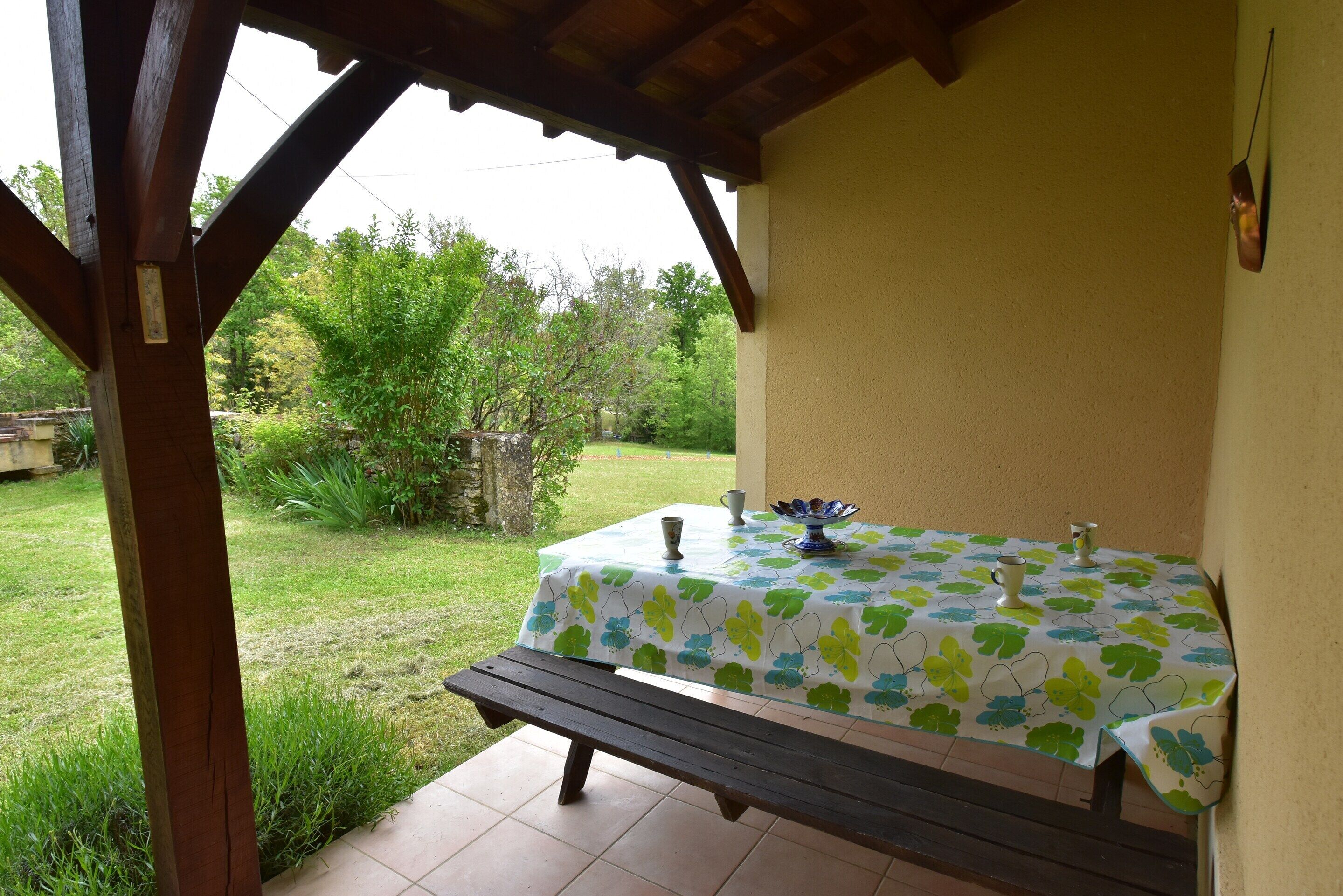A covered patio with a table set for two, a bench, and a view of a garden.