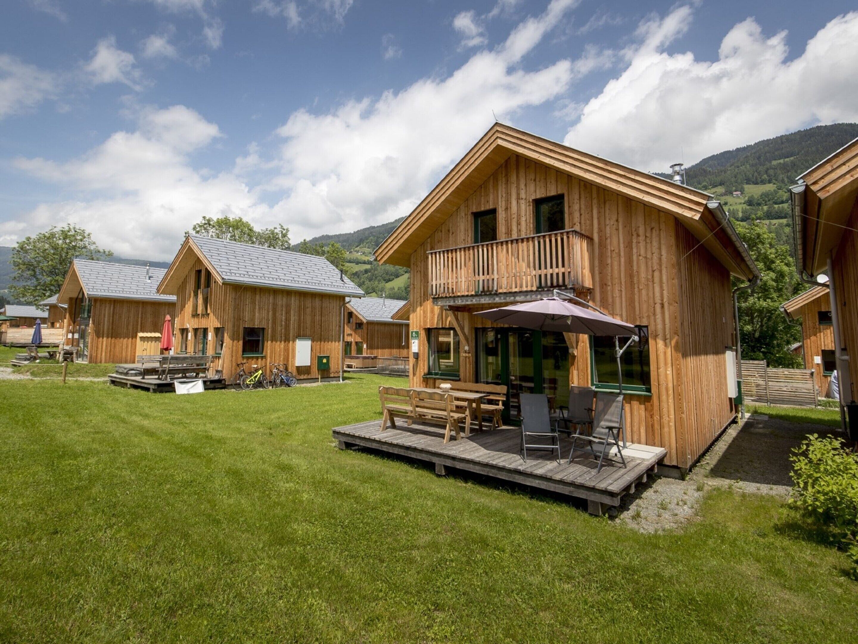 En træhytte med balkon, en terrasse med siddepladser og et bjerg i baggrunden.