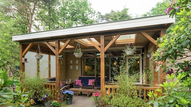 A covered patio with wooden furniture, hanging baskets, and potted plants. - Holiday Home in Arrien Near Beerze Nature (Arriën)