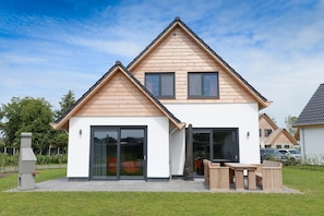 A modern two-story house with a sloped roof, large sliding glass doors, and a patio with outdoor furniture. - Villa Within National Park (De Koog)