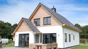 A modern two-story house with a sloped roof, large windows, and a patio with a wooden table and benches. - Villa Within National Park (De Koog)