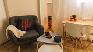 Un espacio acogedor con un sillón oscuro, una mesita blanca pequeña y una mesa de comedor con sillas. En la mesa de comedor hay una vela decorativa y un frutero.