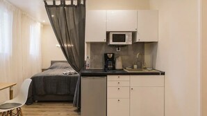 Una habitación compacta con cama, una kitchenette pequeña que incluye microondas, cafetera y lavamanos. Cuenta con una silla y una mesita.