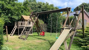 A wooden swing set with a slide, a ladder, and a small playhouse in a grassy backyard.