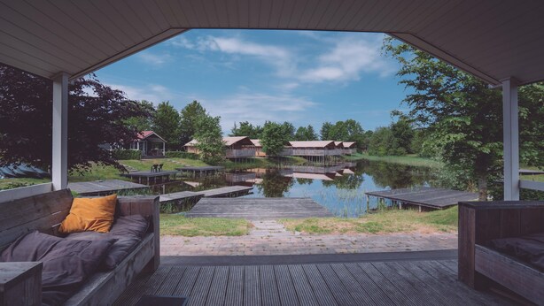 A covered outdoor seating area with wooden benches and a view of a lake and buildings in the background. - Pondside Stay w/ Kitchen (Bant)