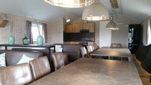 A modern kitchen with wooden cabinets and a black stove, a dining area with a long table and leather chairs, and a large window with curtains. - Pondside Stay w/ Kitchen (Bant)