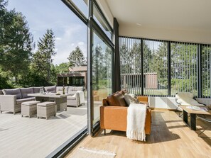A modern living room with a large glass door leading to a deck, furnished with sofas, a coffee table, and a fireplace. - Modern Forest Chalet (Enter)