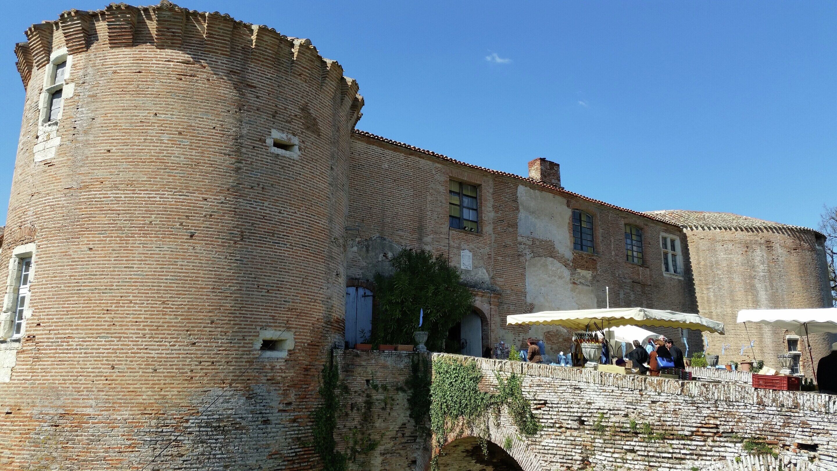 A historic brick building with a large tower, a bridge, and a market area with umbrellas.