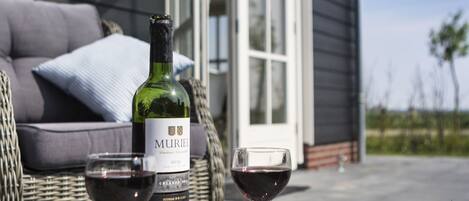 A Muriel wine bottle and two glasses of red wine on a glass table outside a house with a wicker chair and a grey couch.