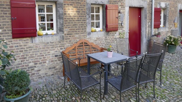A cobblestone courtyard with a wooden table and metal chairs, a brick building with red shutters, and potted plants. - Located Near Maastricht Towards the Belgium Border (Eijsden)
