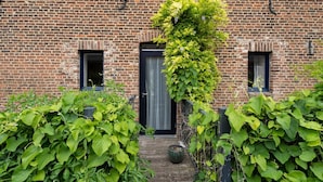 A brick building with a wooden front door, a small window on the left, and a large window on the right. There is a green plant growing over the door and a potted plant on the ground. - Located Near Maastricht Towards the Belgium Border (Eijsden)