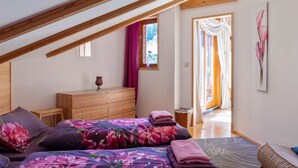 Quarto com teto inclinado, vigas de madeira, cama com roupa de cama floral, penteadeira e janela com cortinas.