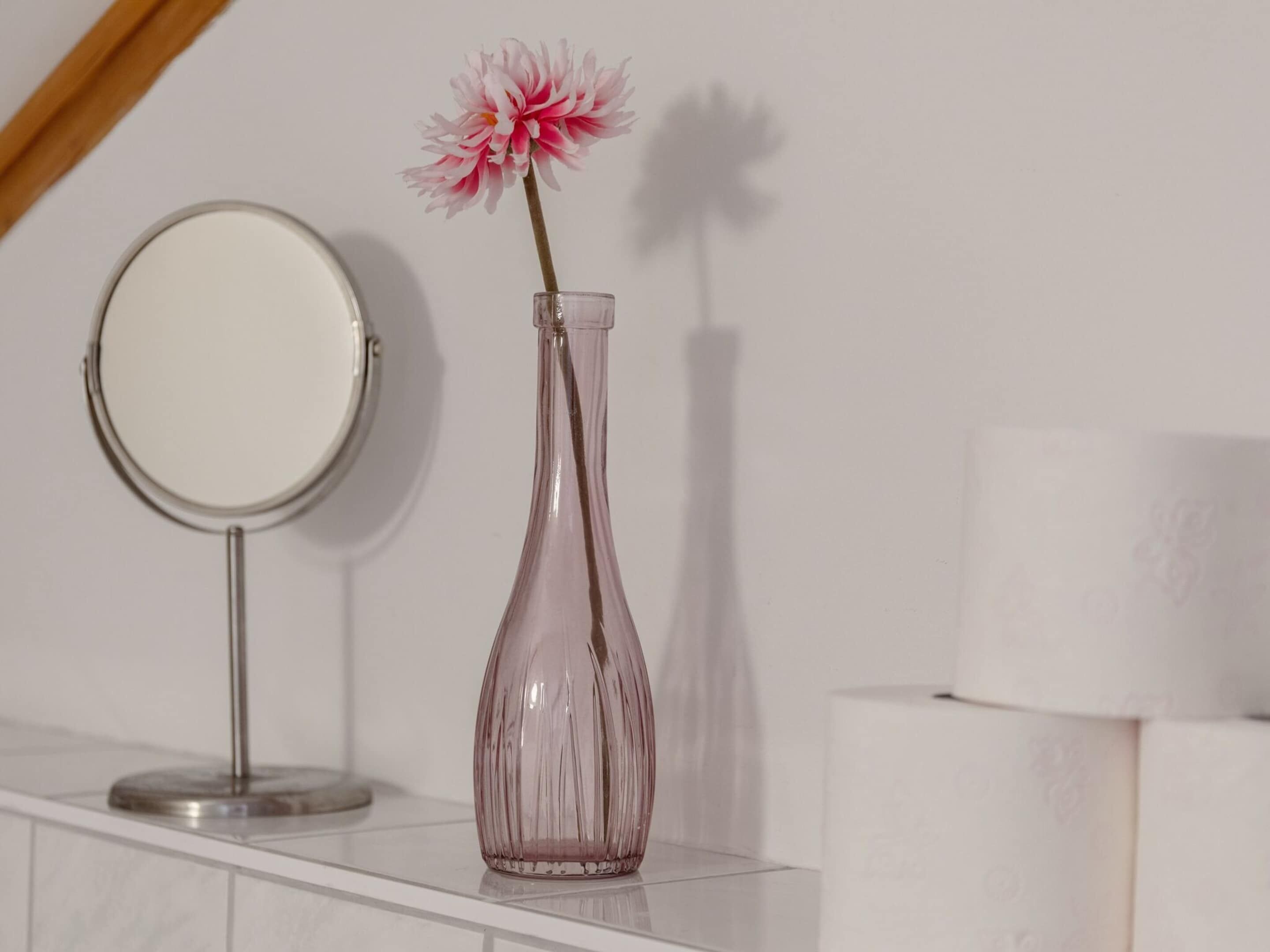 A bathroom vanity with a round mirror, a pink vase with a single flower, and a white towel.