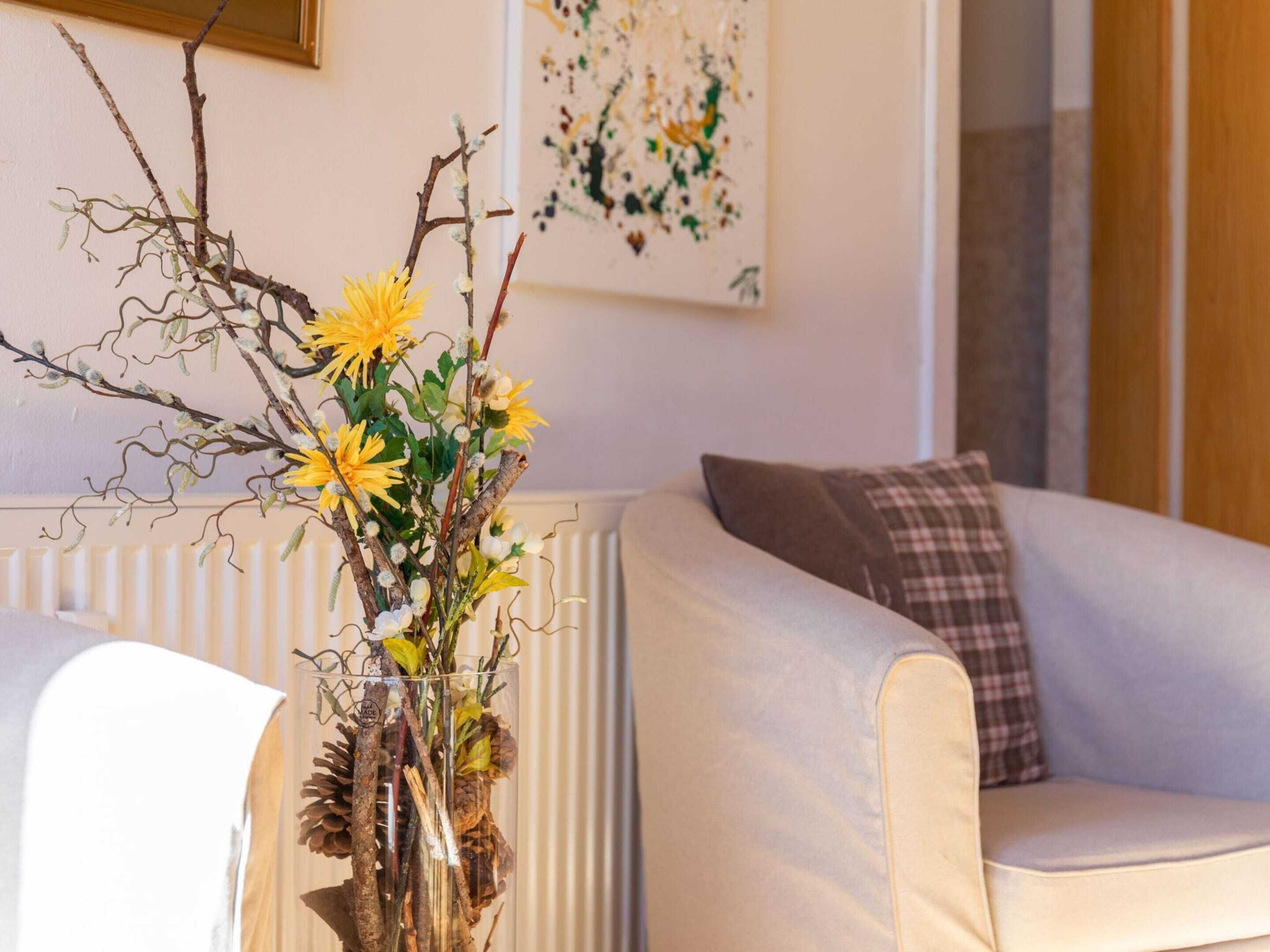 A living room with a white sofa, a glass vase with branches and yellow flowers, and a painting on the wall.