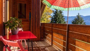 Uma cabana de madeira com varanda, uma mesa vermelha, uma cadeira, um vaso de planta e um guarda-chuva listrado.
