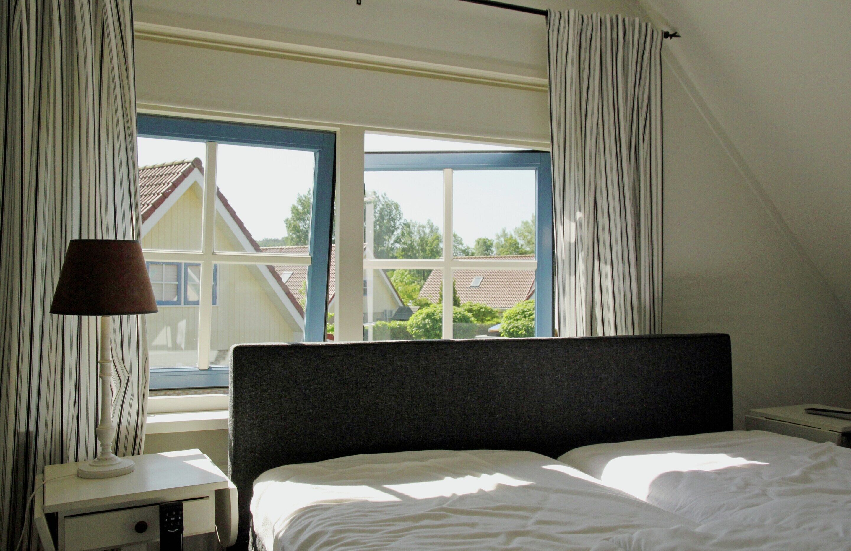 Un dormitorio con cama, mesita de noche, lámpara y ventana con cortinas.