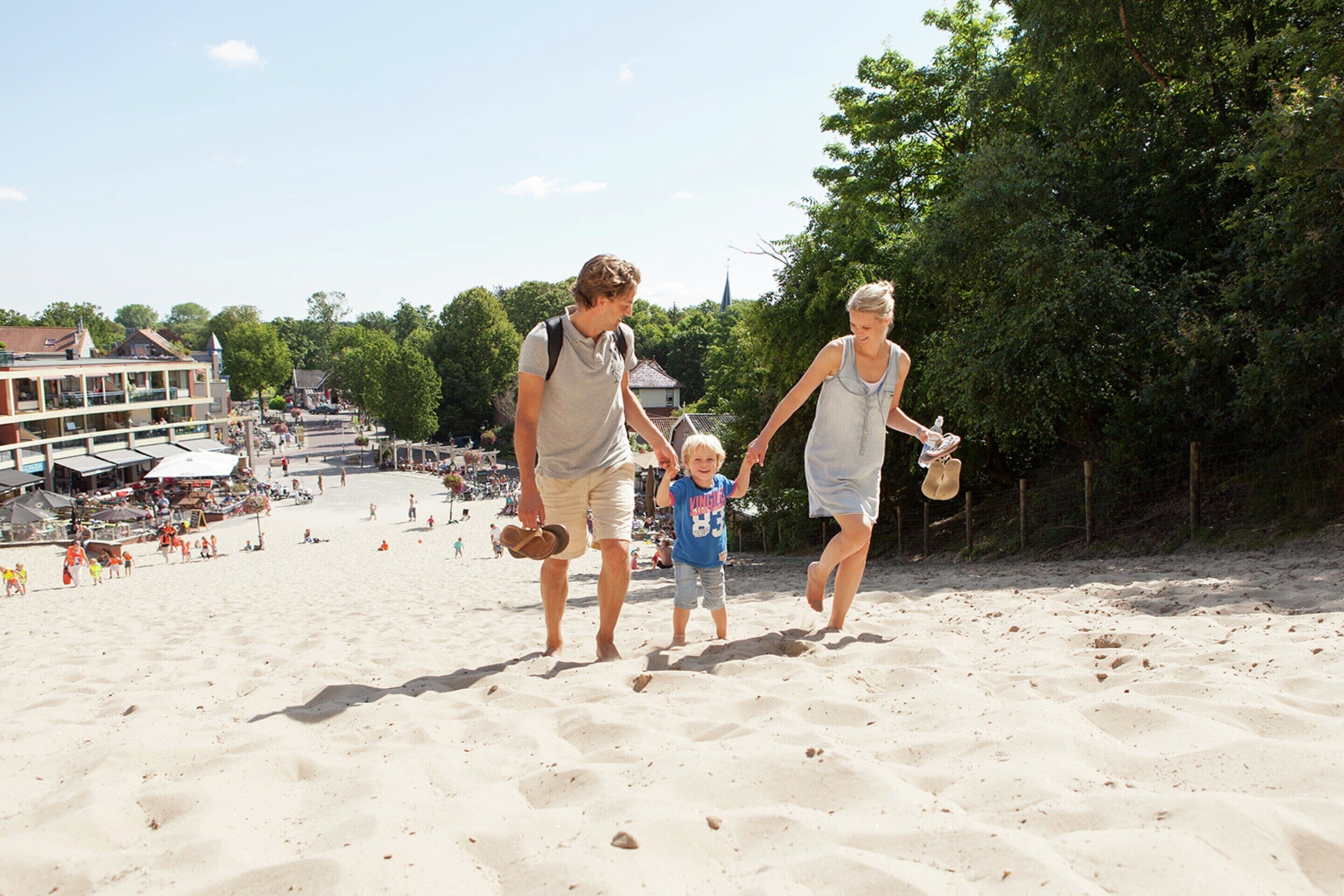 Une plage de sable où l’on voit des gens se promener, un immeuble à étages et des arbres en arrière-plan.